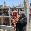 Woman seen from waist up holds a toddler wrapped in a towel while standing in front of bombed-out buildings