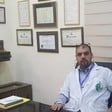 Man in medical uniform sits at desk with framed diplomas on wall behind him