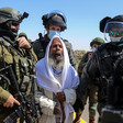 An older man clutches a white shawl worn over his head while soldiers stand around him