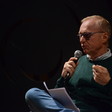 A man in glasses speaks into a microphone with a paper in his lap 