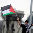 A Palestinian boy wearing a kuffiyeh holds a Palestinian flag in front of a military watch tower