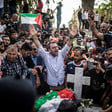 Man holds his hands above his head while standing in crowd around grave with Palestine flag on it