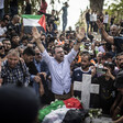 Man holds his hands above his head while standing in crowd around grave with Palestine flag on it