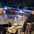 Man and woman sit on chairs on a paved area surrounded by trees in the evening