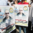 People hold two identical posters side by side showing the face of a bald man wearing sunglasses 