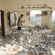 Man holding mobile phone stands in rubble of destroyed home with holes in walls showing view of hill