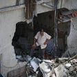 A man looking down with his arms akimbo stands on rubble in a hole in the wall of a bombed-out building