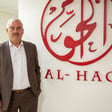 Shawan Jabarin, wearing sports coat and collared shirt, stands in front of wall bearing a large reproduction of Al-Haq's calligraphic logo