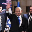 Benjamin Netanyahu, wearing suit and tie, waves while standing in front of Israeli flags