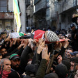 Crowd of men, some carrying rifles, stand in street while body shrouded in Palestine flag is carried on stretcher