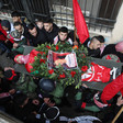 Men and boys carry the body of a boy wrapped in a flag and covered in a wreath and a poster on a stretcher 