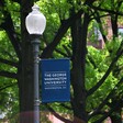 A blue banner with the name of the university hangs on a black lamppost, with trees in the background 