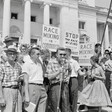 Man speaking at microphone in front of crowd at the Arkansas State Capitol protesting school integration with signs reading Race mixing is Communism and Stop the race mixing