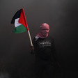 A person with their face covered emerges through dark smoke with the Palestinian flag