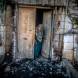 A women peers out from a metal door to an area covered in burned debris