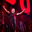 Roger Waters, dressed in a black t-shirt and black jeans and holding a microphone, raises his arms and smiles onstage