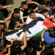 A crowd of men carries the body of a man covered in a flag with a rifle placed on top 