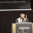 Fatima Mohammed, dressed in a graduation cap and gown, and wearing traditional Palestinian scarves,  stands at a podium.