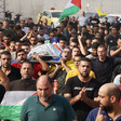 Body shrouded in kuffiyeh and Palestine flag is carried on stretcher by crowd of men, some carrying Palestine and Fatah flags