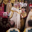 Welby in gold-trimmed bishop's robes places ornate crown on head of seated, robed monarch Charles as others look on