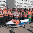 Men wearing orange safety vests stand with their arms crossed in front of their stomachs while standing in front of a shrouded body on a stretcher with a civil defense jacket placed on his chest