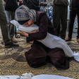 A young woman cries while holding her arms tight around a shrouded body and kneeling on the ground