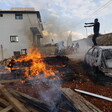 A fire blazes beside a burnt car amidst the destruction caused by a nearby building.