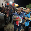 Wael Dahdouh carries a toddler while wearing a flak jacket and as other journalists stand around him, many with cameras