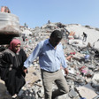 A woman wearing a red head scarf and black clothes and a man wearing a blue shirt and beige trousers walk amid the remains of a building that has been destroyed 