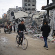 A man on a bicycle rides on a street interspersed with other people with bombed-out buildings on either side