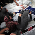 Women sit next to and lean over several bodies shrouded in white sheets