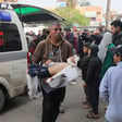 A man walks in front of an ambulance, carrying an injured toddler in his arms amidst a large crowd of people 