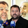 Saleh al-Arouri, seen from chest up, stands between news media microphones and banner showing Hamas leaders Abdel Aziz al-Rantisi and Sheikh Ahmed Yassin