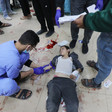 A man wearing medical scrubs holds a syringe above the arm of a boy laying on tiled floor smeared with blood