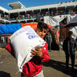 A Palestinian youth carries a bag of UNRWA flour with two youths and an UNRWA school in background
