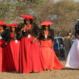 A group of women in bright flowing dresses and hats walk amid trees 