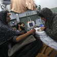 A woman with an injured face and a man laying on a hospital bed reach towards the shrouded body of baby held by a woman who is lifting fabric to expose the child's face