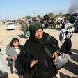 An older woman carries a small purse as she walks on sandy road with two young children carrying blankets close behind her and other people carrying their belongings traveling by foot, donkey cart and car at a distance behind her