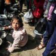 A girl sits on the ground with several empty tin pots. Other people are standing behind her in line