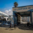 People stand outside gate to UNRWA facility with collapsed structure next to it