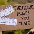 A cardboard protest sign reads "The Hague Awaits You Two" with mock plane tickets for Yoav Gallant and Antony Blinken