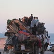 Children sit on top of truck piled high with household objects with sea in background