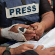 A man wearing a press vest holds the hand of someone wrapped in shroud