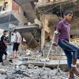 A boy with a bandaged leg uses crutches while walking across rubble in front off bombed-out building