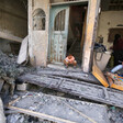 A small child sits in the doorway of a home missing a wall while looking at debris in the street