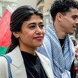 Portrait of the lawmaker Rima Hassan standing in front of a flag and a man wearing a checkered scarf