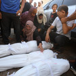 A woman sitting on the ground reaches her arm across three shrouded bodies as other mourners stand nearby