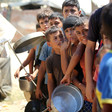 Palestinian boys, some holding pots, stand in a line at a food distribution site.