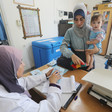 A woman holds a baby who looks towards the camera while standing in front of health clinic examination table