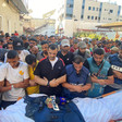 A crowd of mourners pray over a body, wrapped in a white shroud, with a damaged press jacket and an Al Jazeera microphone placed on top.